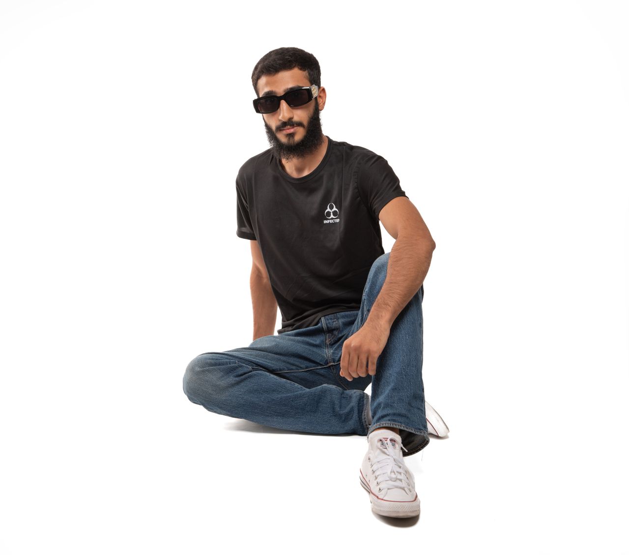 Home A stylish young man in a black shirt and jeans, seated against a white background.